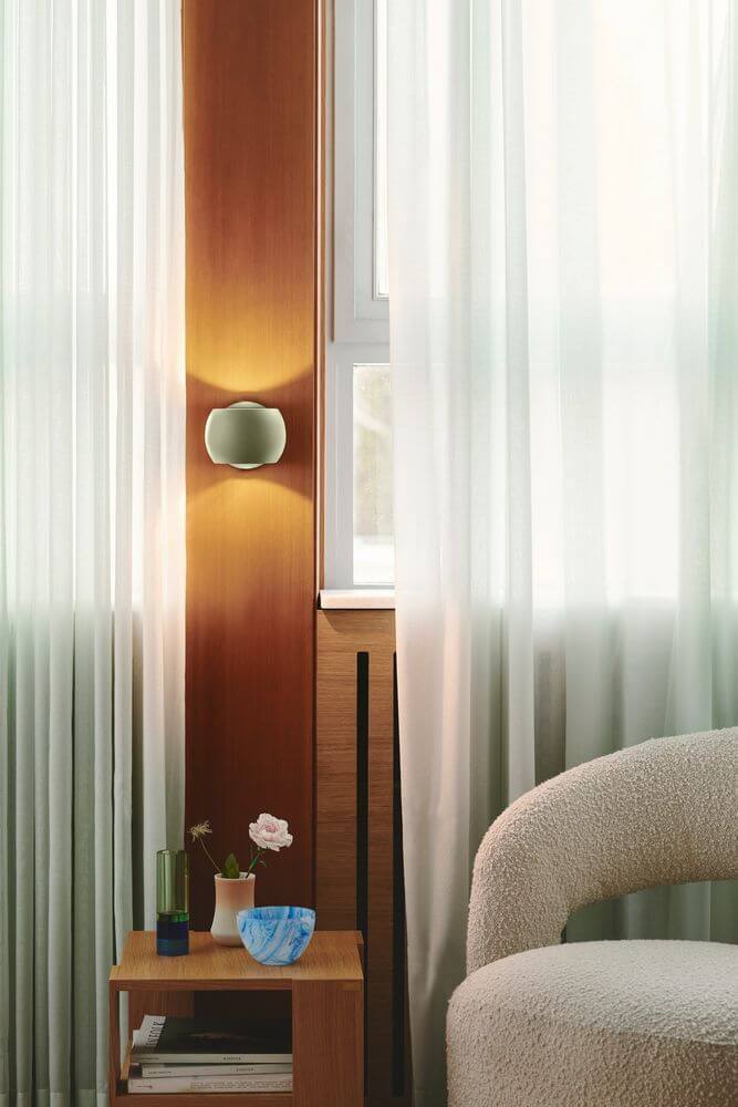 Wandleuchte Belir an einer Wand neben einem gemütlichen Sofa mit Vorhängen und dekorativen Elementen.
