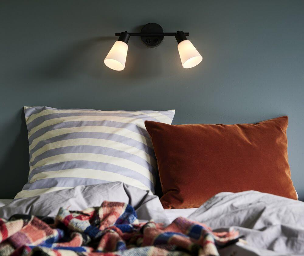 Wandleuchte Cole 2-Spot über einem Bett mit gestreiften und samtigen Kissen.