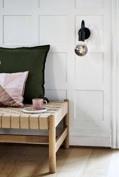 Applique Paco sur un mur au-dessus d’un lit avec des coussins et une table avec une tasse.