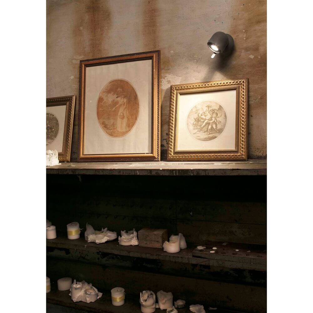 Wandleuchte STONE-1 Grau beleuchtet Kunstwerke in einem rustikalen Raum mit Kerzen auf dem Regal.