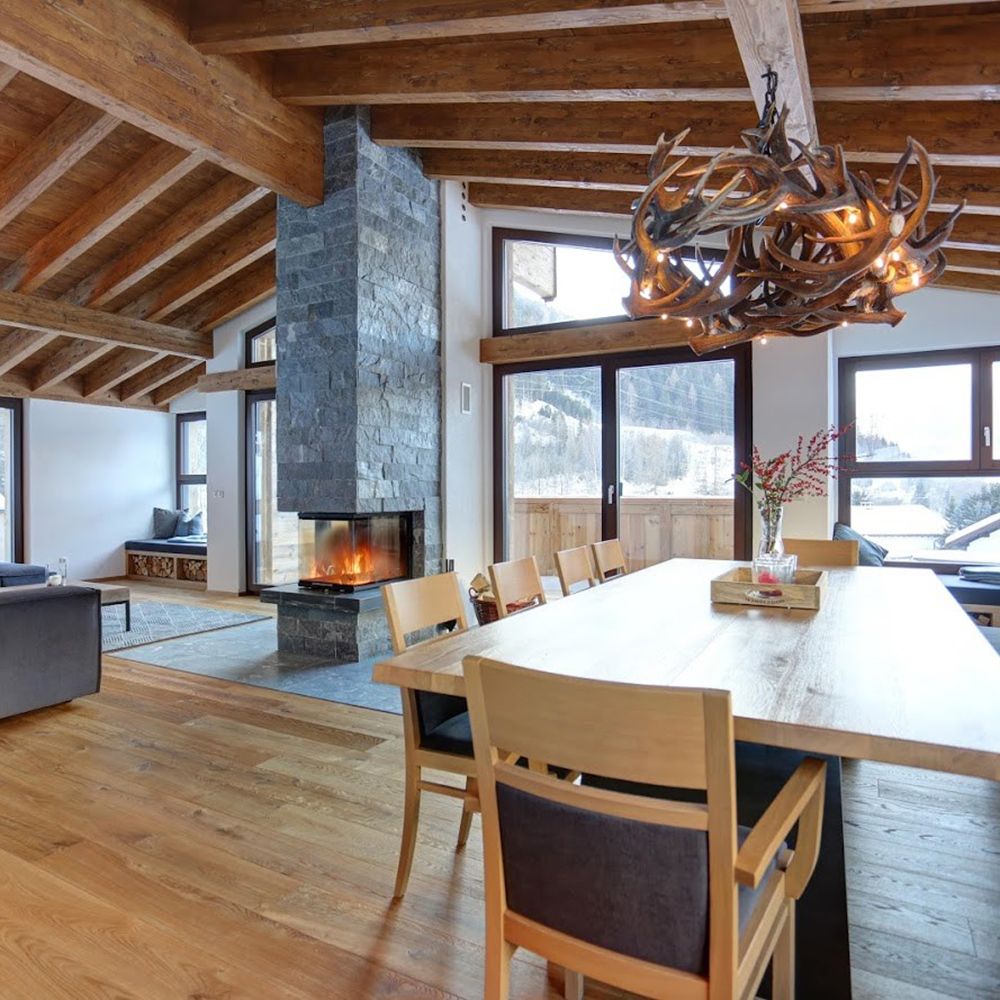 Gemütlicher Wohnbereich mit XXL Rothirsch LED Geweihlampe und Kamin, Holzdecke und großen Fenstern für viel Licht.