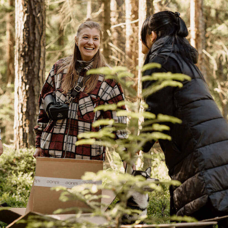 Fröhliche Frauen im Wald, die mit Paketen und Pflanzen interagieren, umgeben von Bäumen und Natur.