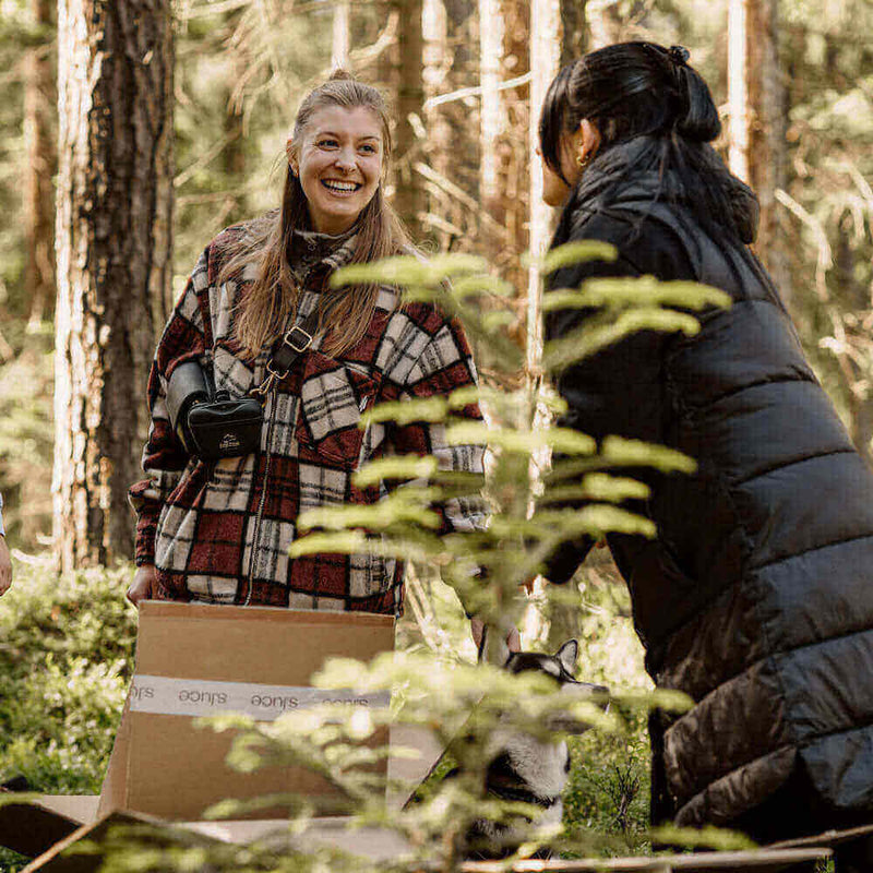 Fröhliche Frauen im Wald, die mit Paketen und Pflanzen interagieren, umgeben von Bäumen und Natur.