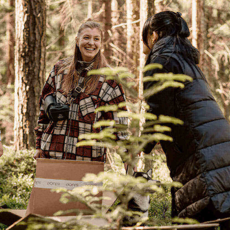 Fröhliche Frauen im Wald, die mit Paketen und Pflanzen interagieren, umgeben von Bäumen und Natur.