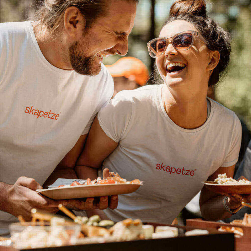 Lachendes Paar in weißen Shirts mit "skapetze" beim Genießen von Essen im Freien.