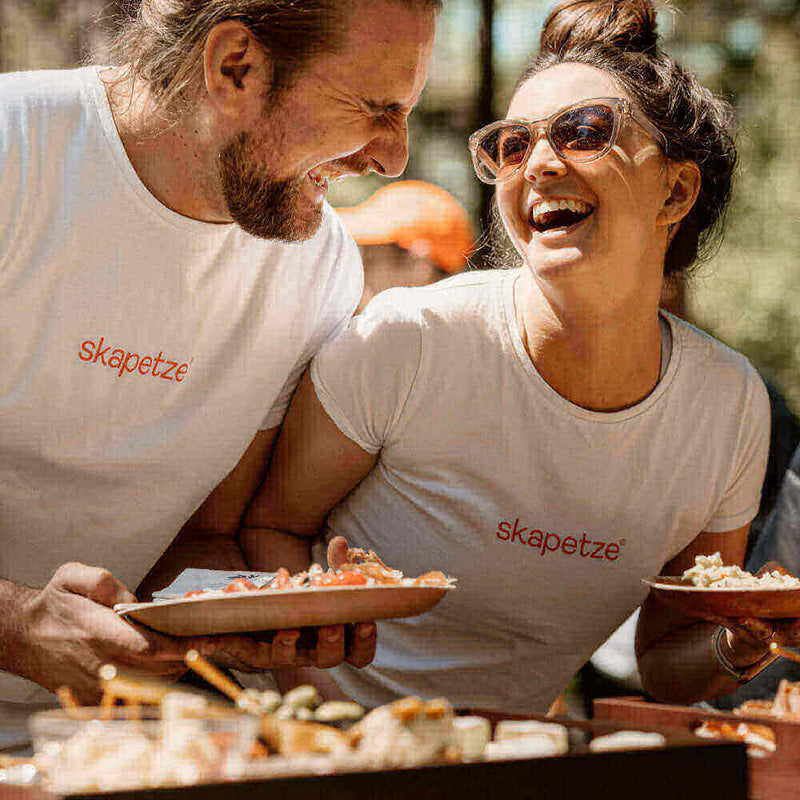 Lachendes Paar in weißen Shirts mit "skapetze" beim Genießen von Essen im Freien.