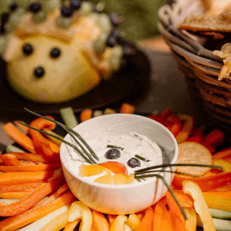 Bunte Gemüseschale mit Dip, dekorativ angerichtet mit Paprika und einem lustigen Obstgesicht im Hintergrund.