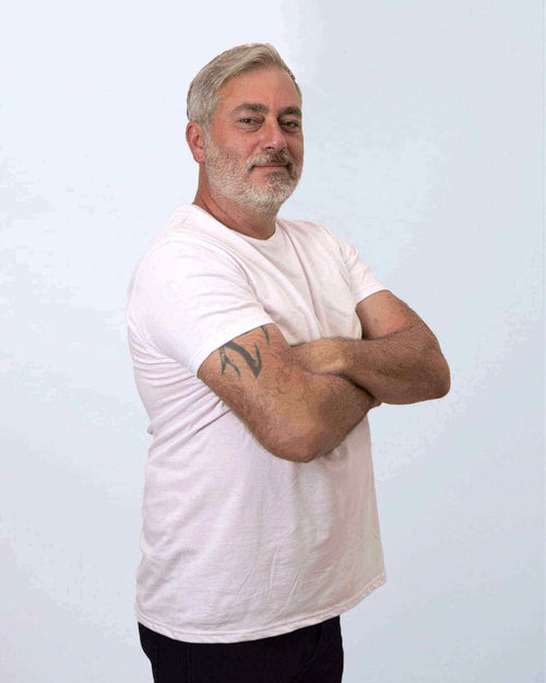 Mann mit kurzem Haar und Bart, der in einem weißen T-Shirt mit verschränkten Armen steht.