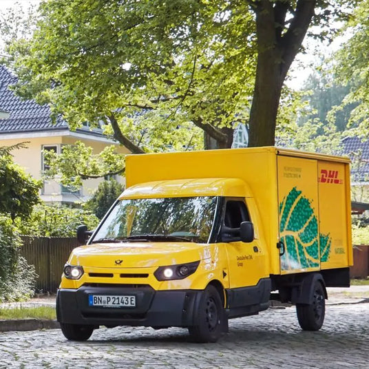 Gelbes DHL Lieferfahrzeug fährt auf einer ruhigen Straße unter Bäumen.
