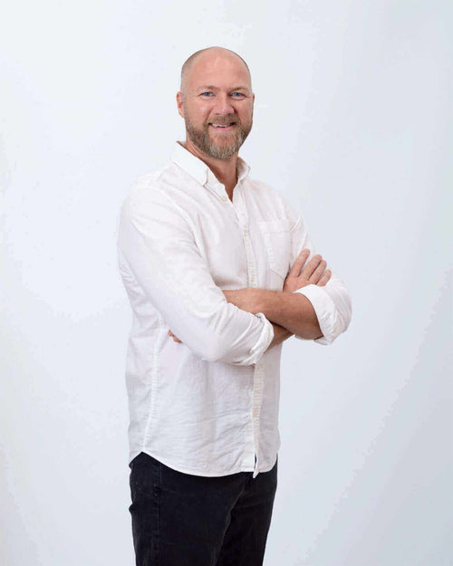 Männlicher Portrait mit verschränkten Armen, lächelnd in einem weißen Hemd vor neutralem Hintergrund.