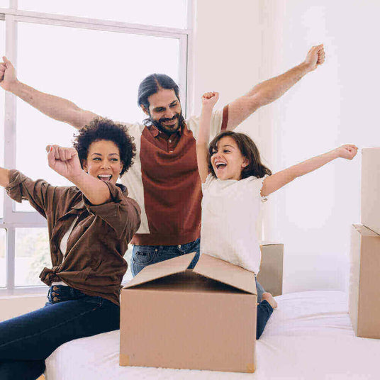 Eine fröhliche Familie mit einem Kind, das in einem neuen Zuhause zusammen feiert, mit Umzugskartons um sie herum.