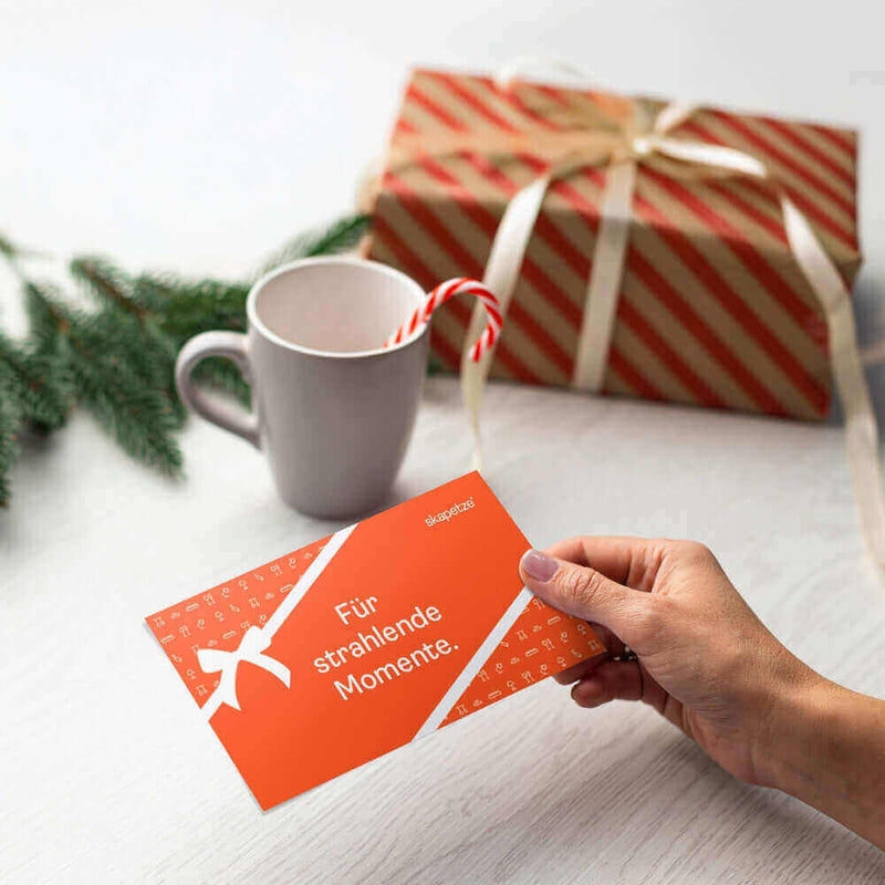 Hand hält eine orangefarbene Karte mit dem Text 'Für strahlende Momente' neben einer Tasse und einem Geschenk.