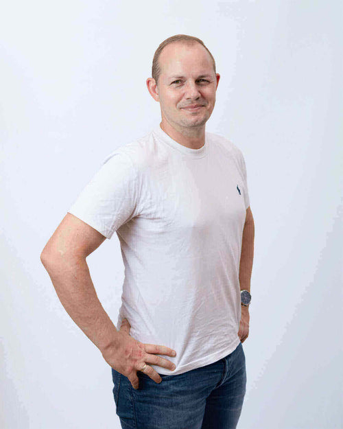 Mann steht mit verschränkten Armen, lächelnd, trägt ein weißes T-Shirt und blaue Jeans.