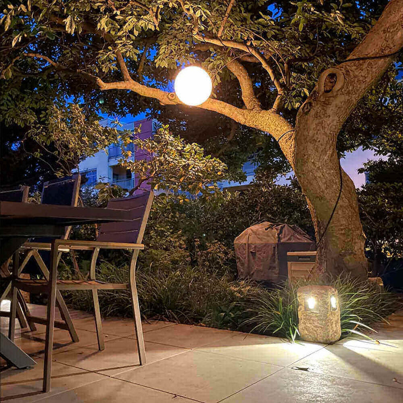 Gemütlicher Garten am Abend mit leuchtender Kugel im Baum und stilvoller Beleuchtung auf dem Tisch.