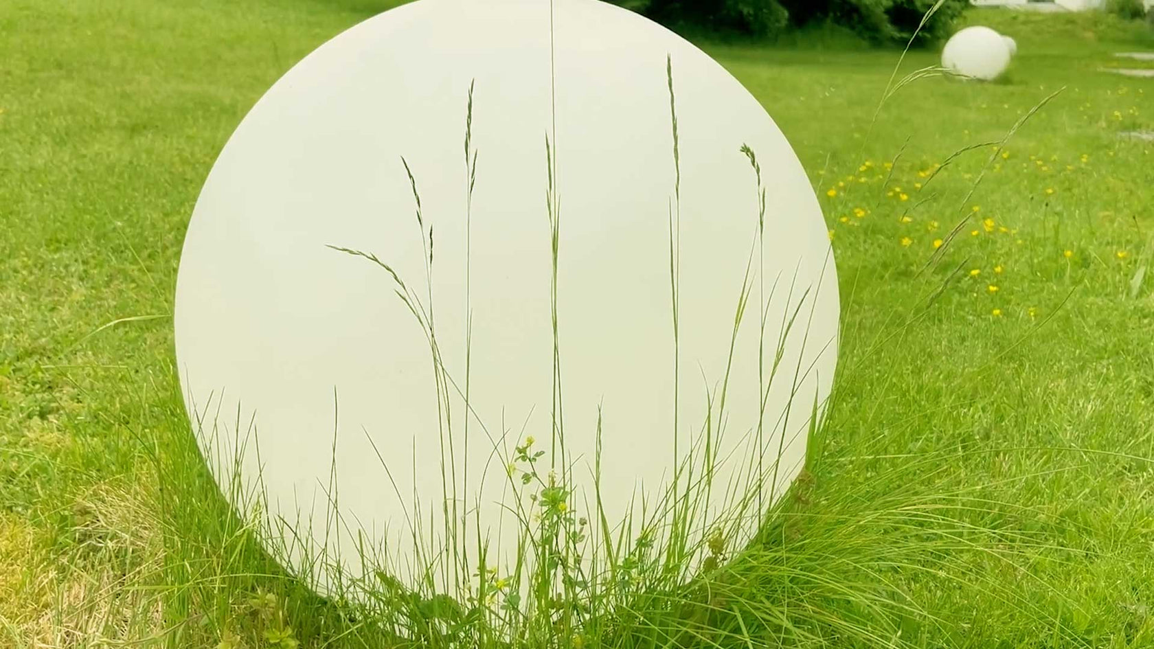 Runde, weiße Form im Grasfeld mit hohen Gräsern und gelben Blumen im Hintergrund.