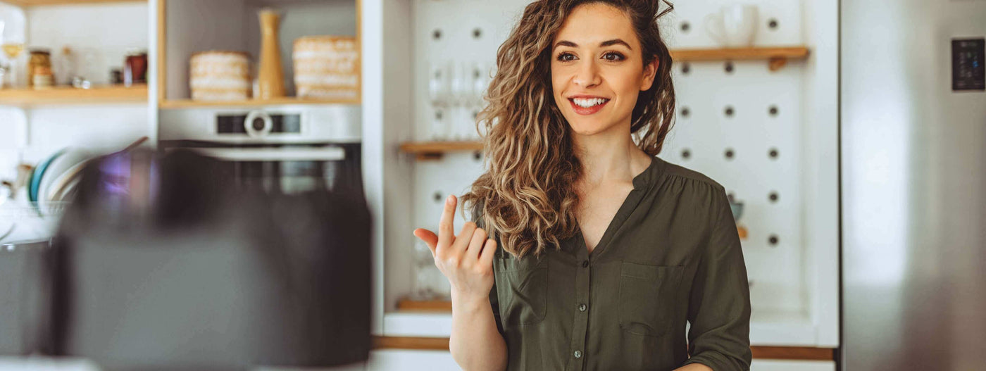 Lächelnde Frau mit lockigem Haar in einer modernen Küche, die eine Kamera anspricht.