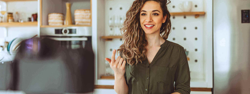 Lächelnde Frau mit lockigem Haar in einer modernen Küche, die eine Kamera anspricht.