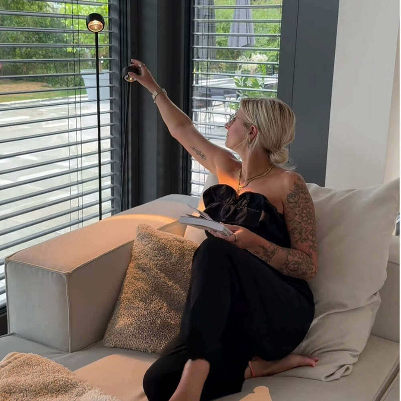 Frau mit tätowierten Armen, die in einem modernen Wohnzimmer Jalousien öffnet, um Licht hereinzulassen.