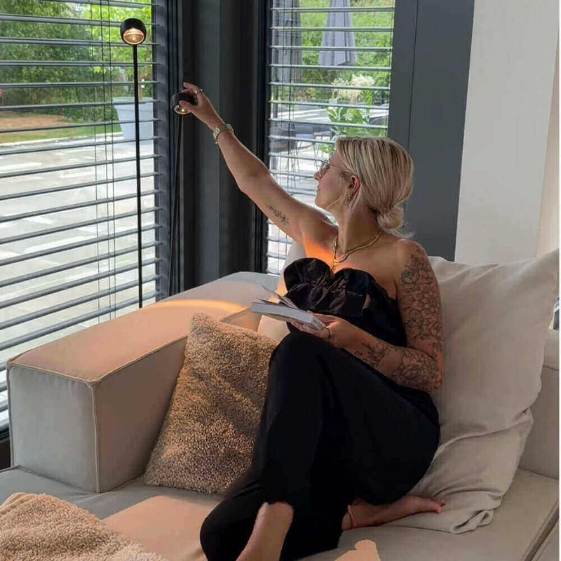 Frau mit tätowierten Armen, die in einem modernen Wohnzimmer Jalousien öffnet, um Licht hereinzulassen.