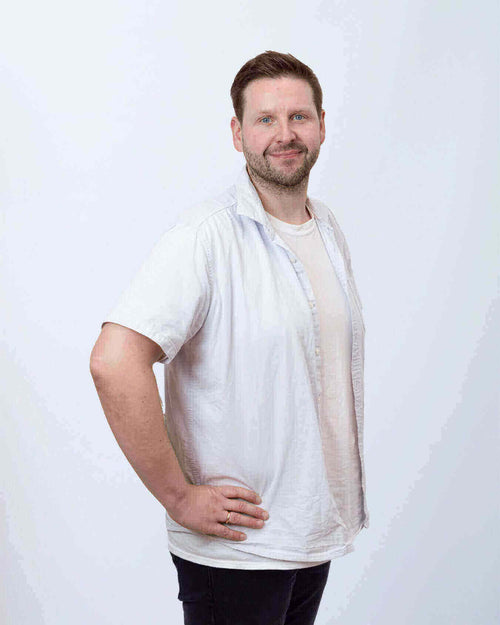 Mann mit hellen Hemd und freundlichem Lächeln, in hellen Farben, stehend vor neutralem Hintergrund.