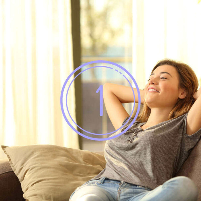 Junge Frau entspannt auf einem Sofa mit hellem Hintergrund und lächelt zufrieden.