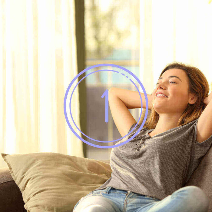 Junge Frau entspannt auf einem Sofa mit hellem Hintergrund und lächelt zufrieden.