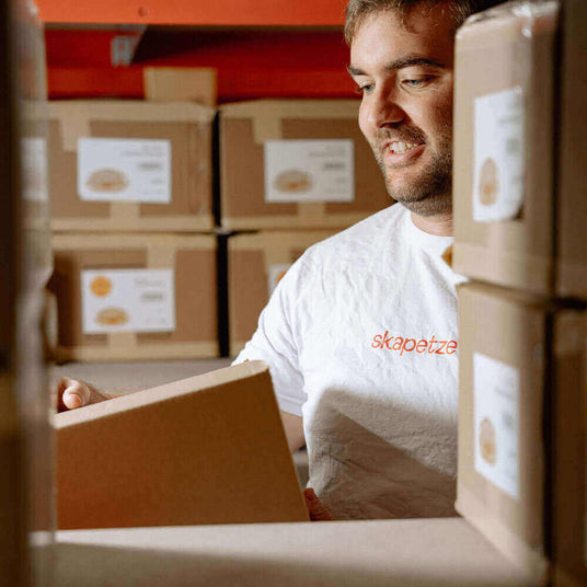Mitarbeiter öffnet eine Box in einem Lager voller Kartons, grinsend mit einem T-Shirt von skapetzer.