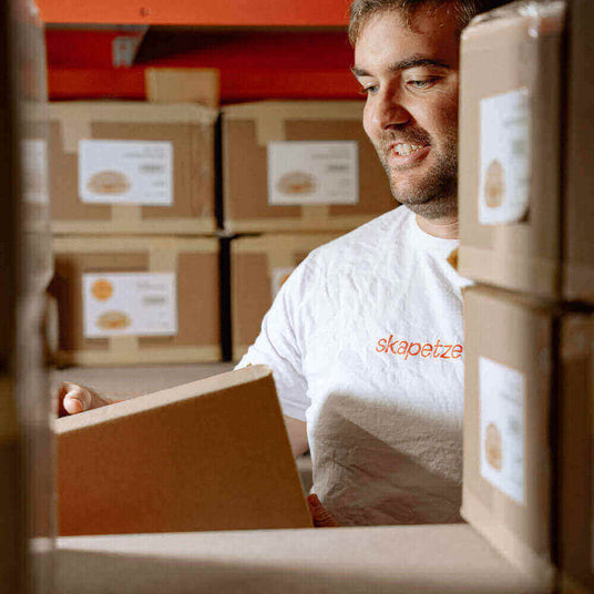 Mitarbeiter öffnet eine Box in einem Lager voller Kartons, grinsend mit einem T-Shirt von skapetzer.