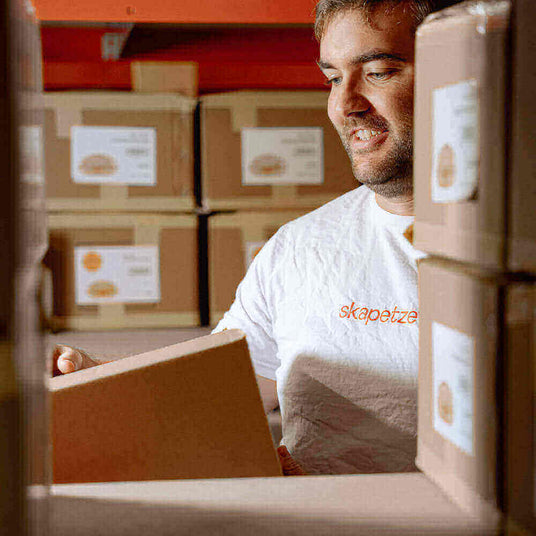 Mitarbeiter öffnet eine Box in einem Lager voller Kartons, grinsend mit einem T-Shirt von skapetzer.