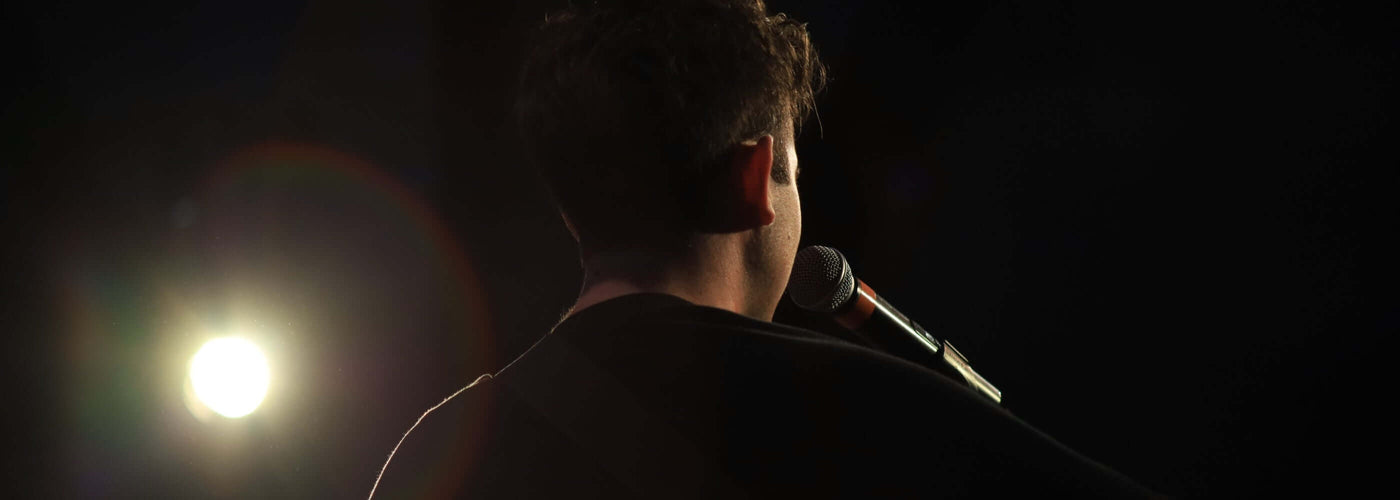 Sänger von hinten mit Mikrofon auf der Bühne, beleuchtet von Scheinwerferlicht.