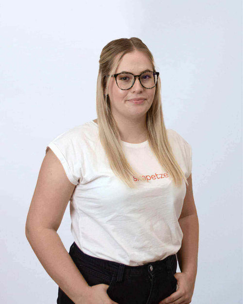 Junge Frau mit Brille in weißem T-Shirt, lächelnd, vor neutralem Hintergrund. Suitable for s.luce und Fashion.