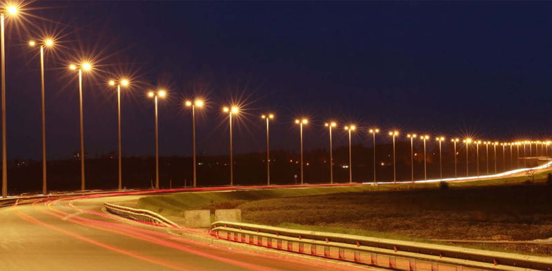 Nachtaufnahme einer beleuchteten Straße mit Straßenlaternen und Fahrzeuglichtern, die vorbeifahren.
