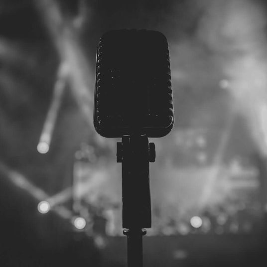Mikrofon im Vordergrund, beleuchtet von Scheinwerfern, auf einer Bühne in Schwarz-Weiß.