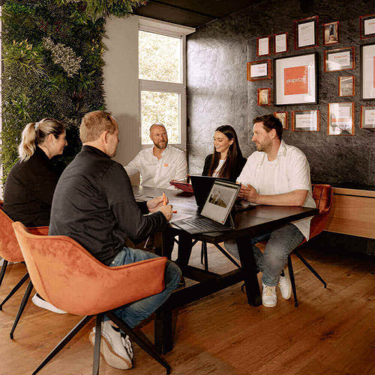 Teammeeting im modernen Konferenzraum mit Laptop und Präsentationen, umgeben von Pflanzen und Auszeichnungen.