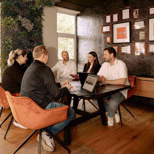 Teammeeting im modernen Konferenzraum mit Laptop und Präsentationen, umgeben von Pflanzen und Auszeichnungen.