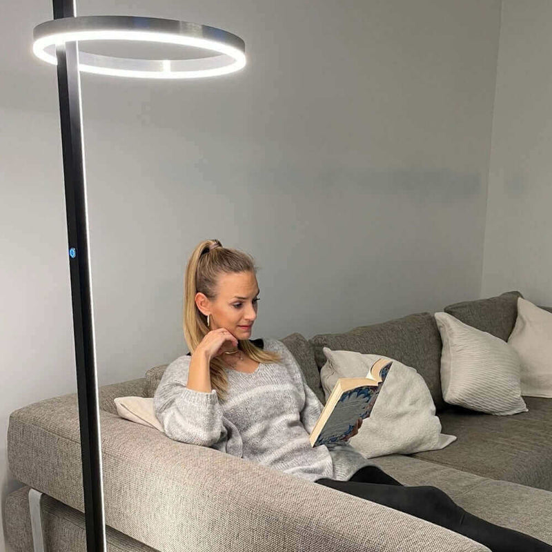 Frau mit Pferdeschwanz liest ein Buch auf einer grauen Couch, beleuchtet von einer modernen Stehlampe.