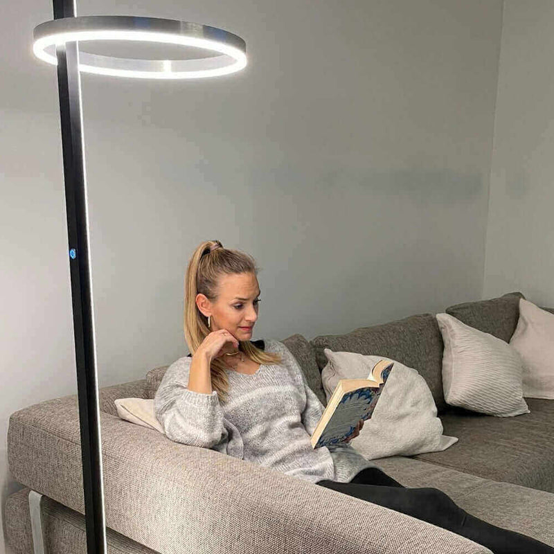 Frau mit Pferdeschwanz liest ein Buch auf einer grauen Couch, beleuchtet von einer modernen Stehlampe.