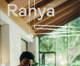 Frau mit Brille arbeitet an einem Tisch in einem modernen Raum mit Holzdecke und Pflanzen im Hintergrund.