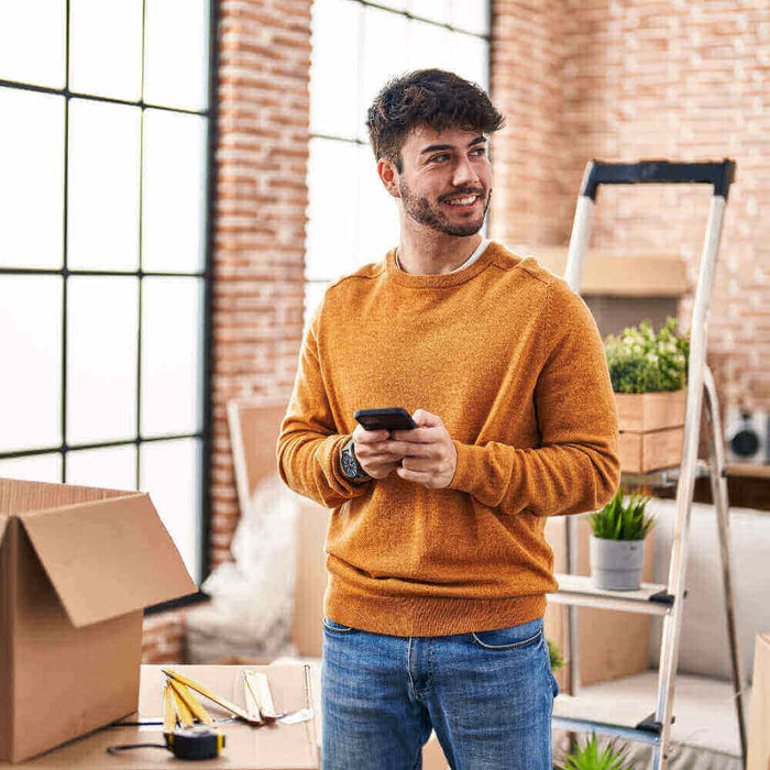 Junger Mann in orangefarbenem Pullover, der in einem gemütlichen Raum mit Umzugskartons und Pflanzen steht.