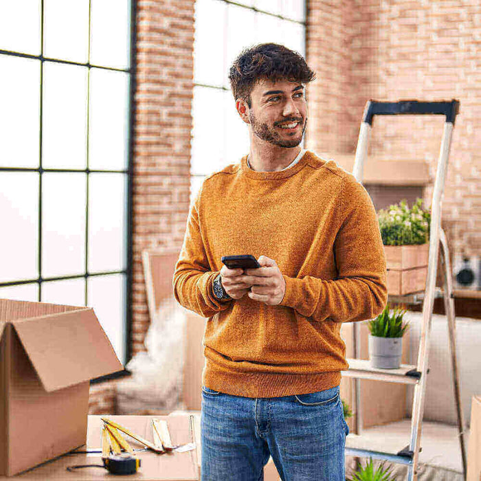 Junger Mann in orangefarbenem Pullover, der in einem gemütlichen Raum mit Umzugskartons und Pflanzen steht.