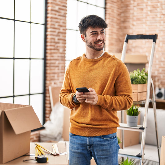 Junger Mann in orangefarbenem Pullover, der in einem gemütlichen Raum mit Umzugskartons und Pflanzen steht.