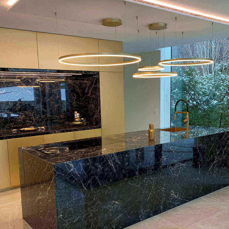 Moderne Küchenansicht mit schwarzen Marmorflächen und eleganten Hängelampen, große Fenster mit Blick ins Grüne.