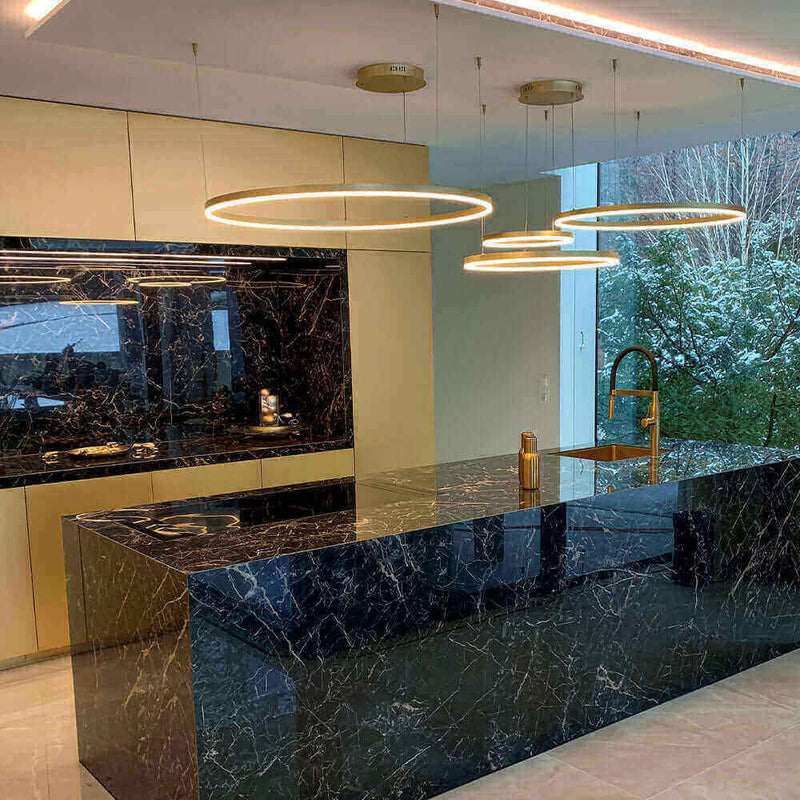 Moderne Küchenansicht mit schwarzen Marmorflächen und eleganten Hängelampen, große Fenster mit Blick ins Grüne.