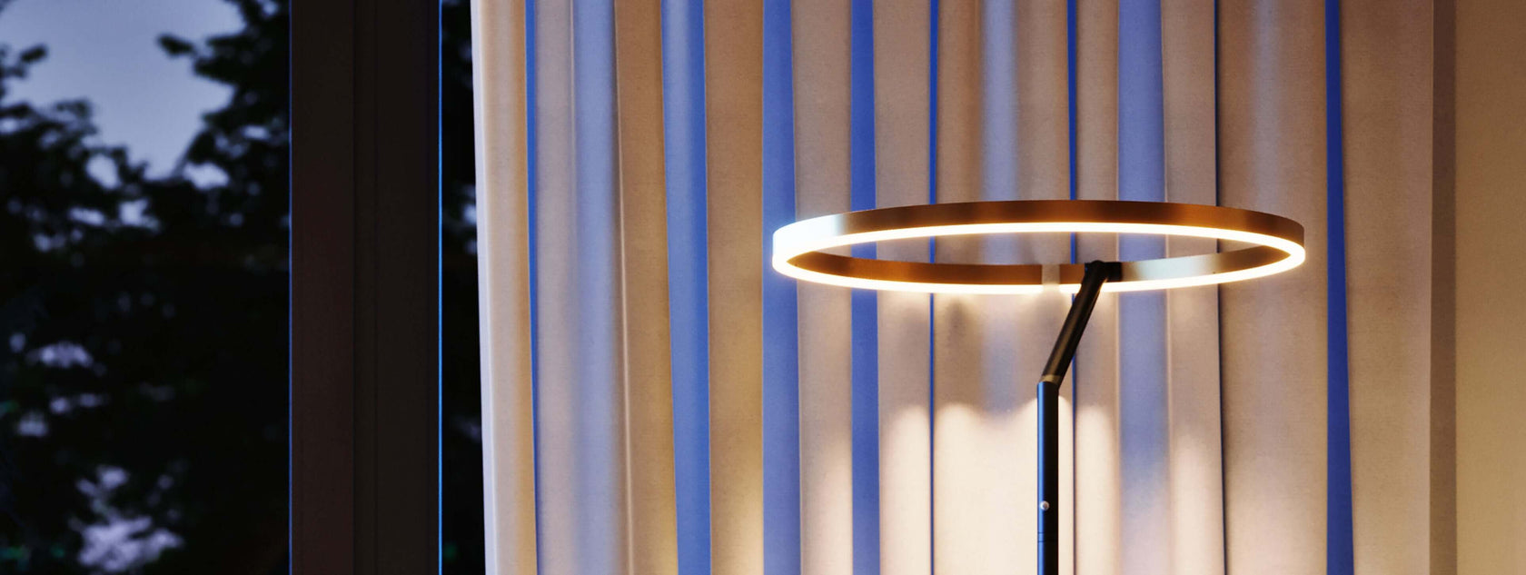 Moderne Lampe mit runder LED-Beleuchtung neben eleganten Vorhängen bei Abendlicht.