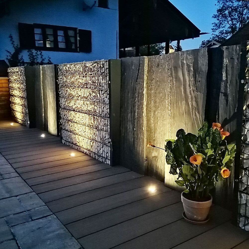 Moderner Außenbereich mit runden Edelstahl Bodeneinbaustrahlern, Steinwand und blühenden Pflanzen bei Nacht.