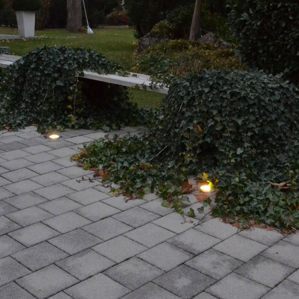 Runder Level Bodeneinbaustrahler in gepflastertem Garten, umgeben von Efeu und sanften Lichtern.