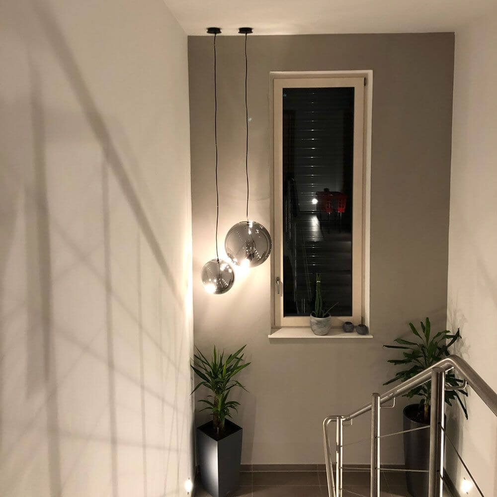 Moderne s.luce Sphere Pendelleuchte in rauchfarbenem Glas, schwebend im Treppenhaus, mit Pflanzen im Hintergrund.