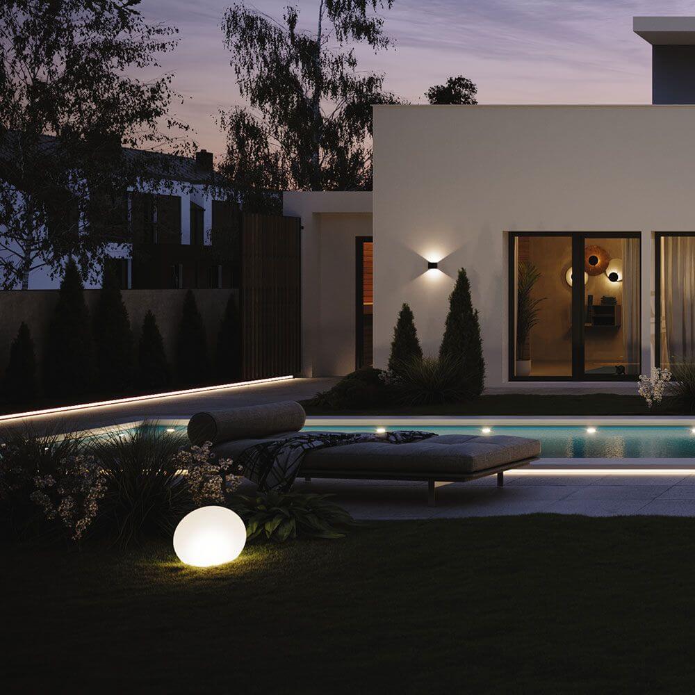 Moderne Ixa LED-Wandleuchte und Balllicht im Garten vor stilvollem modernen Haus bei Dämmerung.