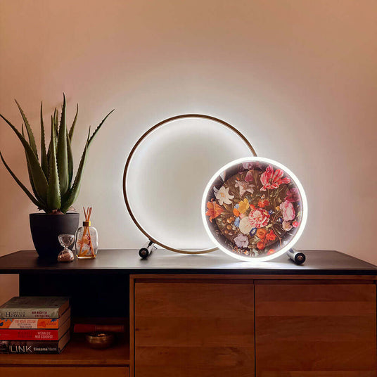 s.luce Leuchte mit floralem Design auf einem Holztisch, daneben eine runde goldene Lampe und Pflanzen.