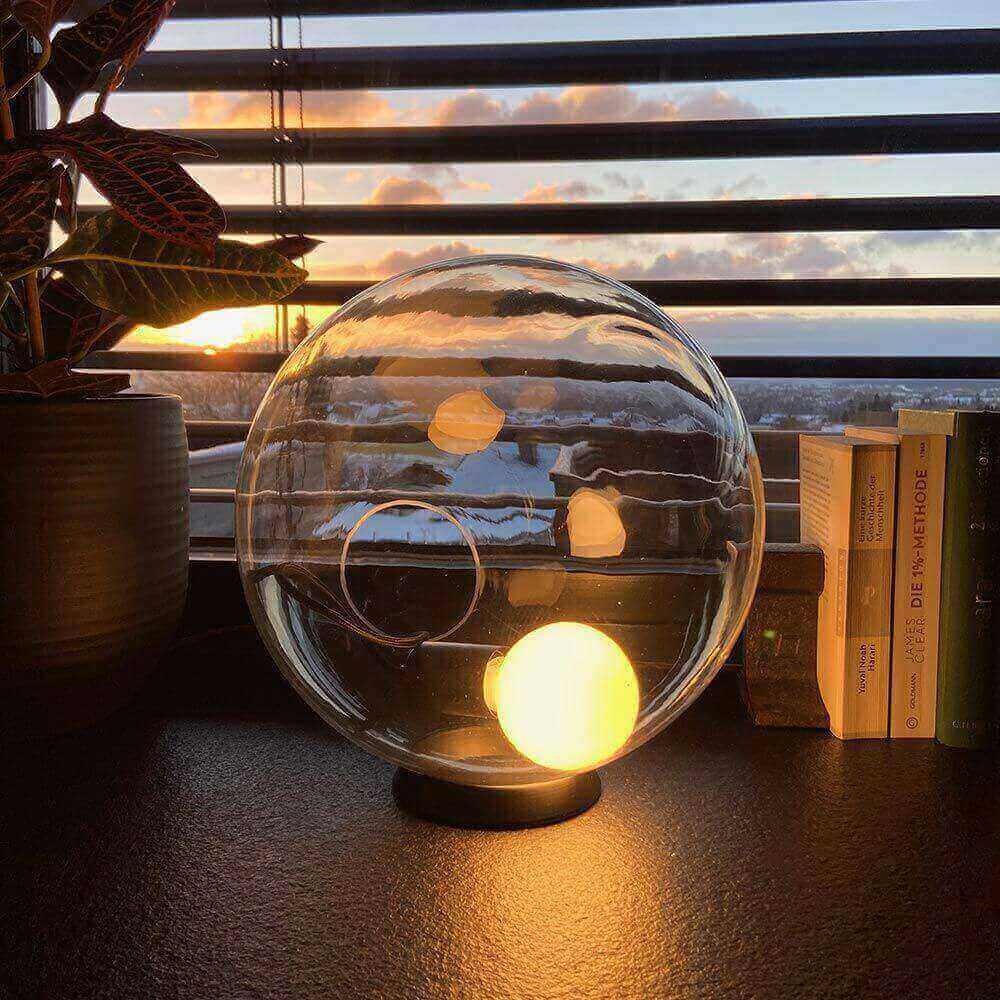 Moderne Tischlampe in einer Glaskugel, mit Sonnenuntergang im Hintergrund und Pflanzen auf dem Tisch.